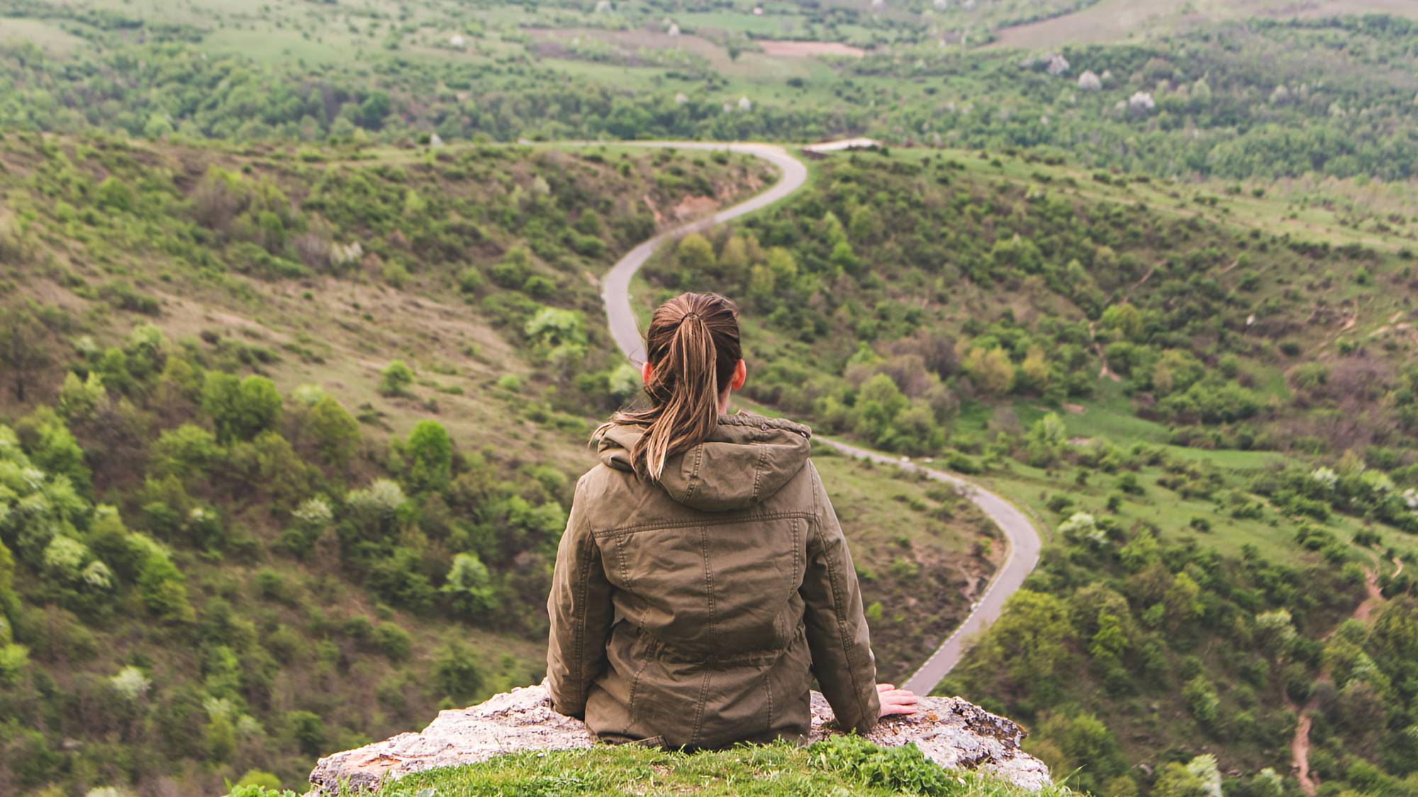 Young girl sitting on a mountaintop overlooking a rural road